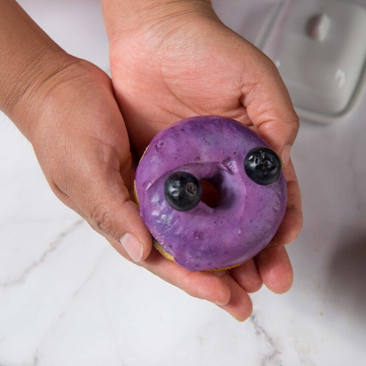 Hand holding a purple FITbelly donut-shaped object with blackberries on a white surface