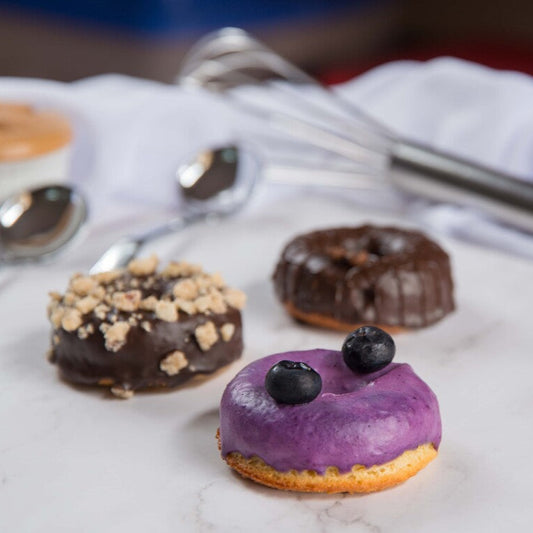 Three FITbelly donuts with different toppings on a marble surface with utensils in the background.
