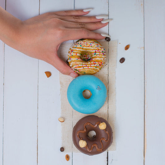 Hand holding a colorful FITbelly donut above three other donuts on a white wooden surface.